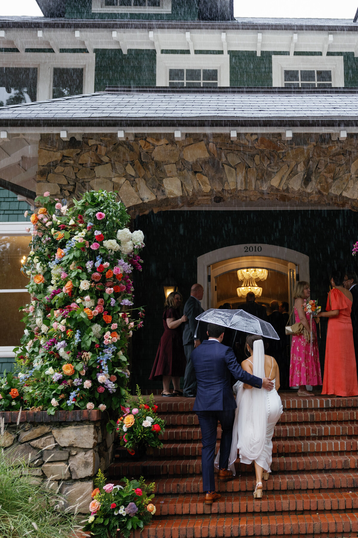 A couple share a clear umbrella as they climb brick steps into a wedding venue in heavy rain, past a tall floral installation