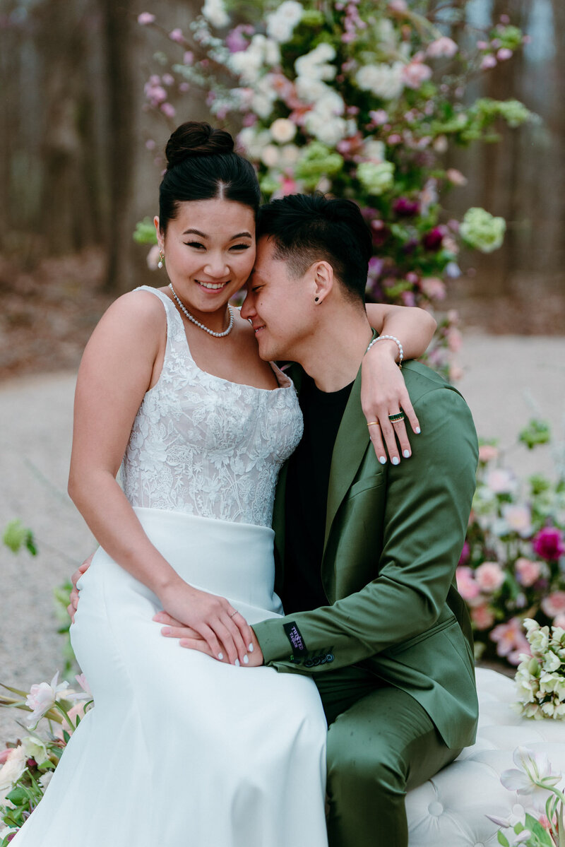 A couple sit close together in front of a tall spring floral installation, one in a lace bodice gown, the other in an olive green suit